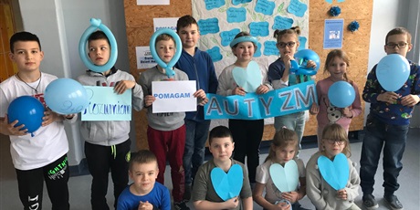 Powiększ grafikę: Uczniowie klasy II, ubrani na niebiesko pozują na tle gazetki o autyzmie, w rękach trzymają niebieskie balony oraz napis AUTYZM